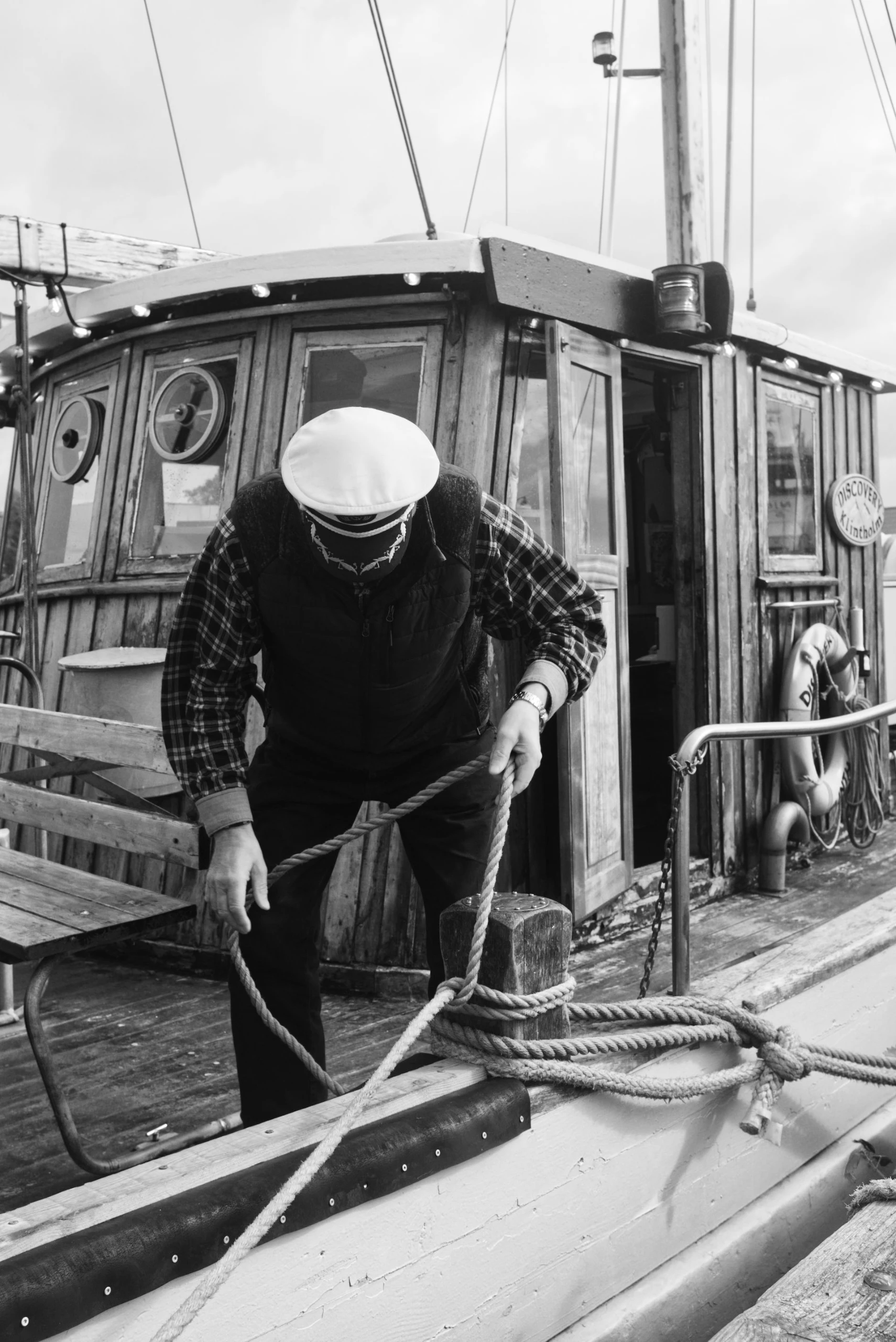 Man met witte pet maakt een touw vast aan een bolder op het dek van een houten schip.